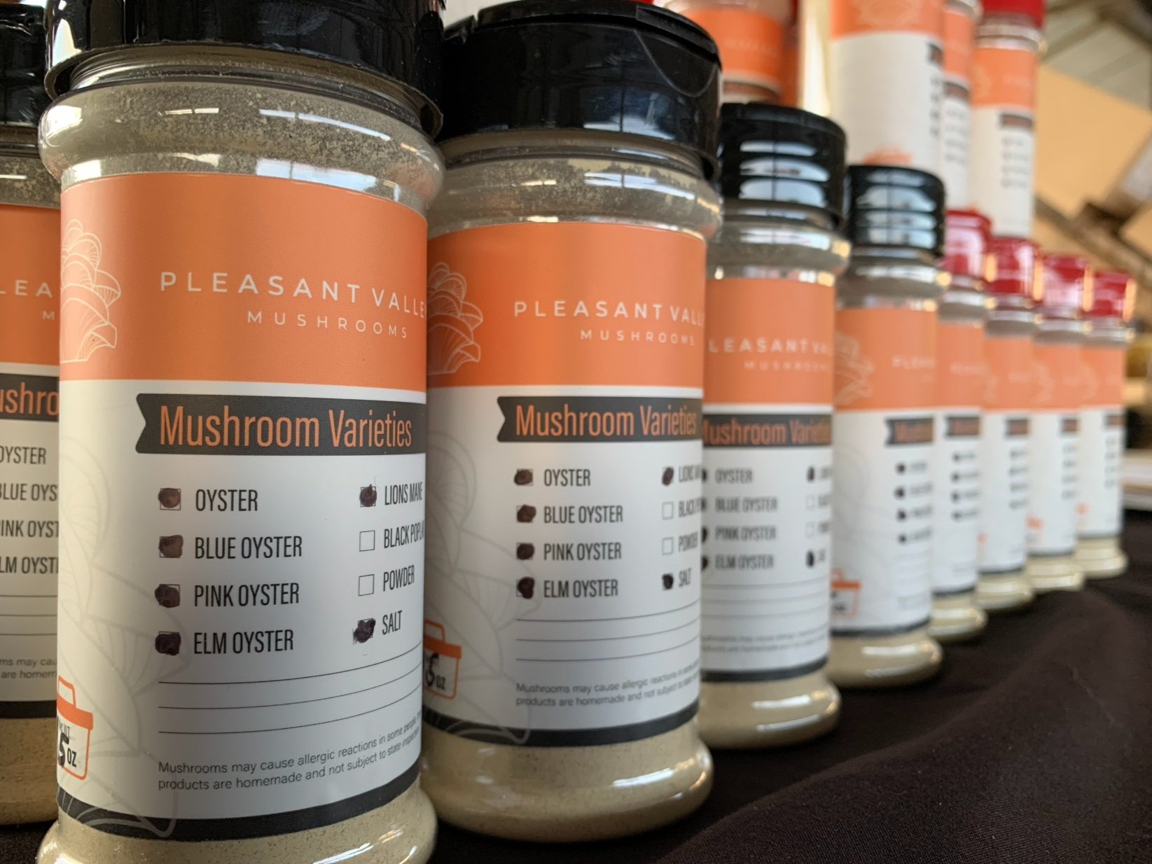 Bottles of Pleasant Valley Mushrooms showcasing various mushroom varieties, including oyster, blue oyster, pink oyster, lion's mane, black pearl, elm oyster, and mushroom powder. The labels provide clear product information, emphasizing quality and variety.